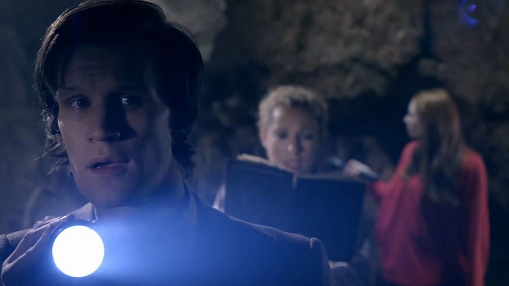 The Doctor in the foreground, with a torch. River is reading from a book in the background