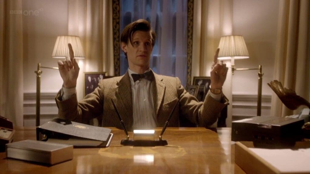 The Doctor behind the desk in the Oval Office, hands up pointing